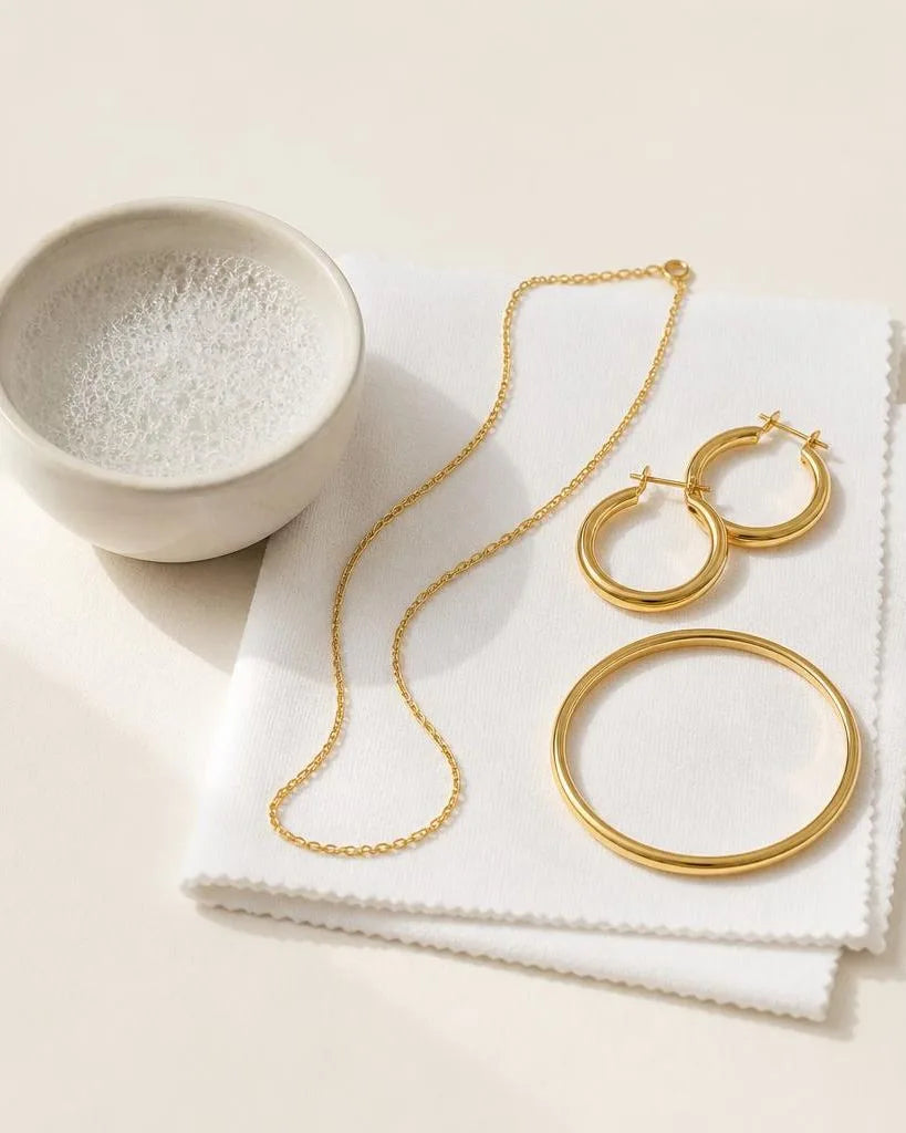 Joyas chapadas en oro colocadas sobre un paño de microfibra junto a un bol de agua jabonosa, listas para una limpieza suave