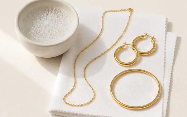 Joyas chapadas en oro colocadas sobre un paño de microfibra junto a un bol de agua jabonosa, listas para una limpieza suave