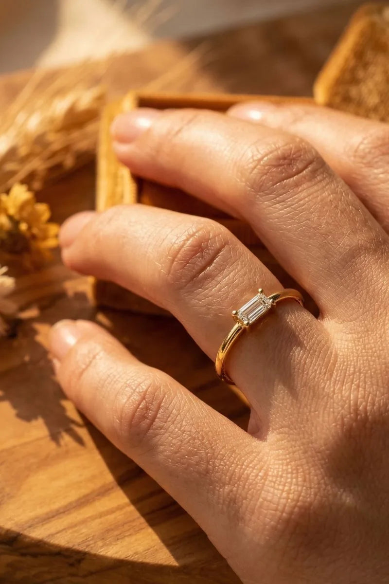Anillo solitario de diamante talla baguette en oro amarillo 18 quilates llevado en el dedo sobre fondo natural por Mayuri